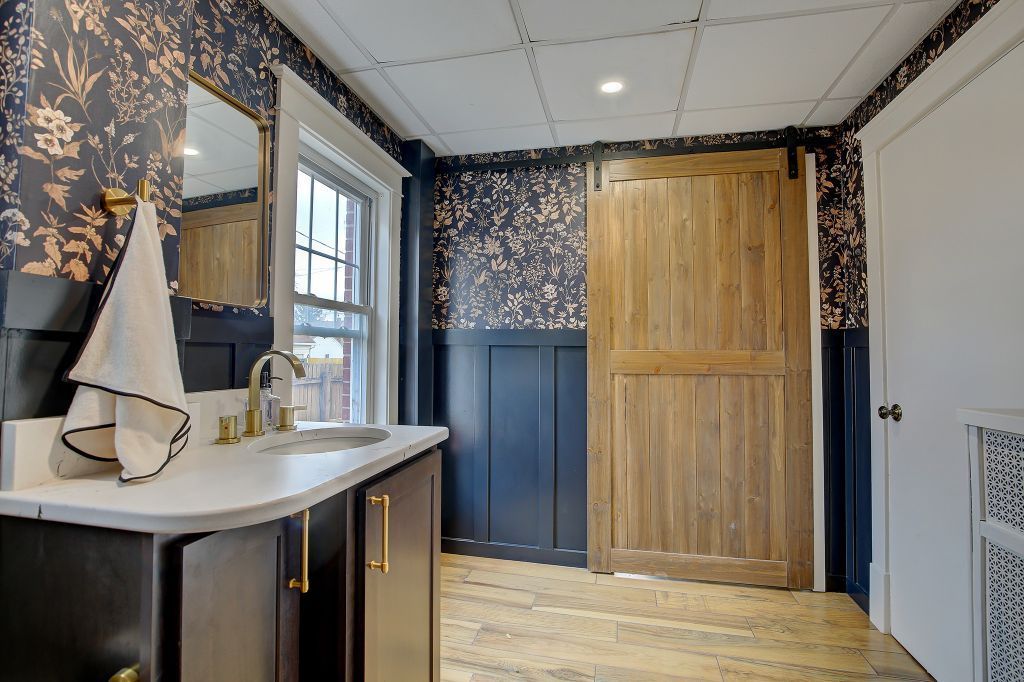 Bathroom with floral wallpaper, vanity sink, wood sliding door, and white cabinet in a bright interior room