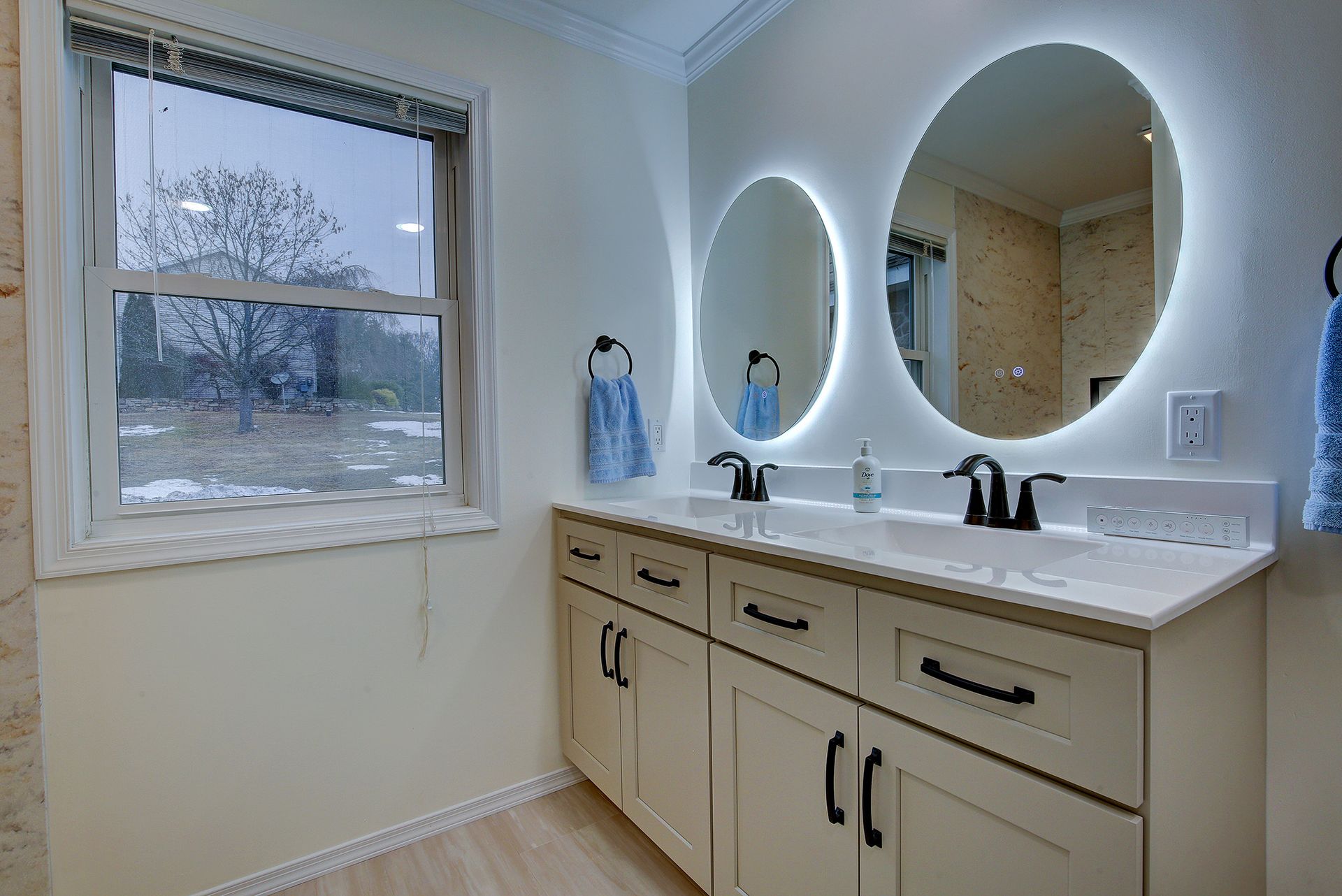 Bright bathroom with double sinks, oval mirrors, and a window overlooking snowy trees