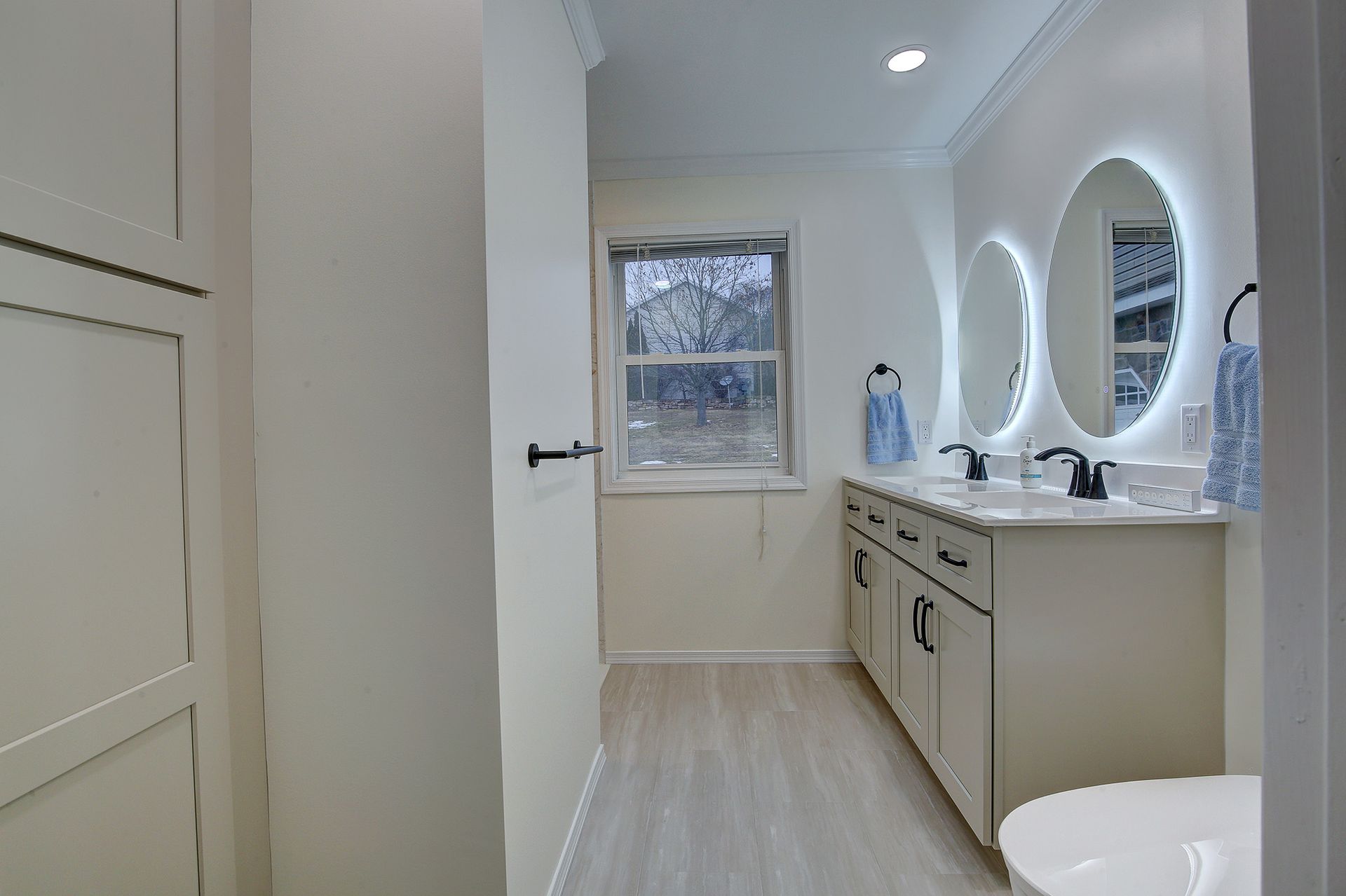 Bright bathroom with vanity, oval mirror, sink, window, and toilet; white walls and light wood flooring