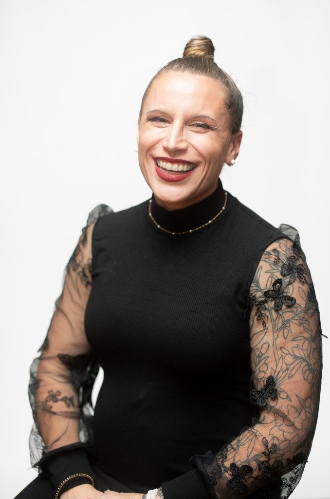 Smiling woman in a black top with sheer lace sleeves, seated against a plain light background