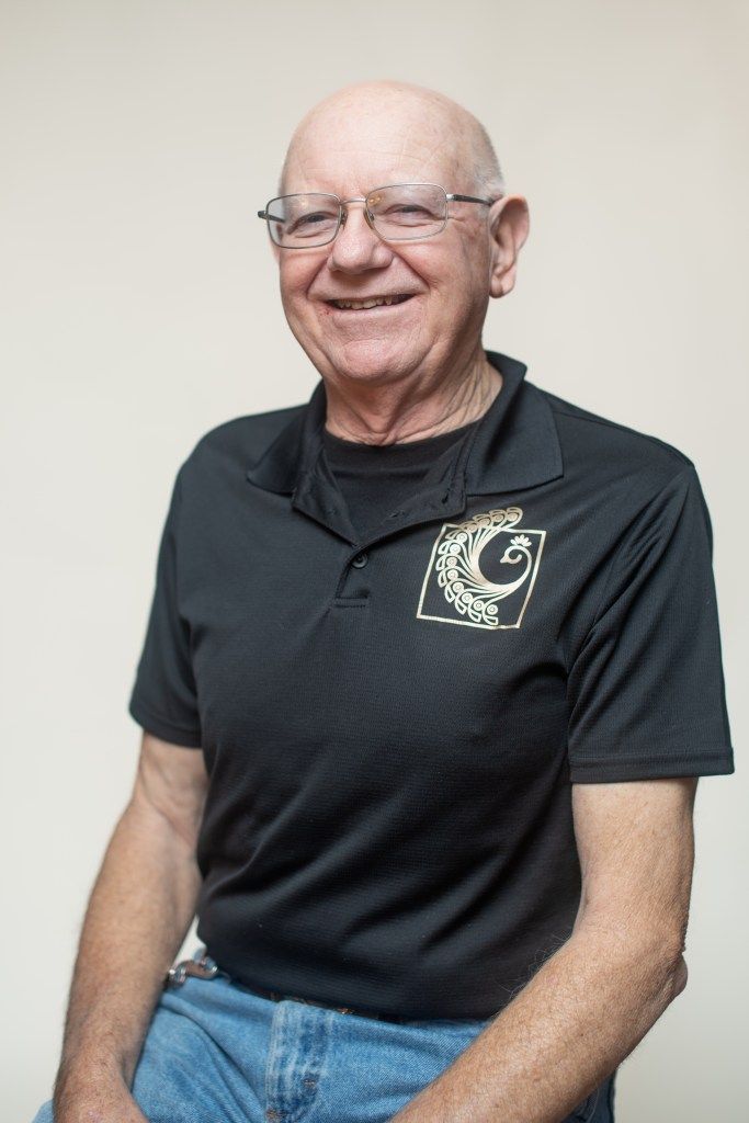 Smiling man in glasses wearing a black polo shirt and jeans, seated against a plain wall.