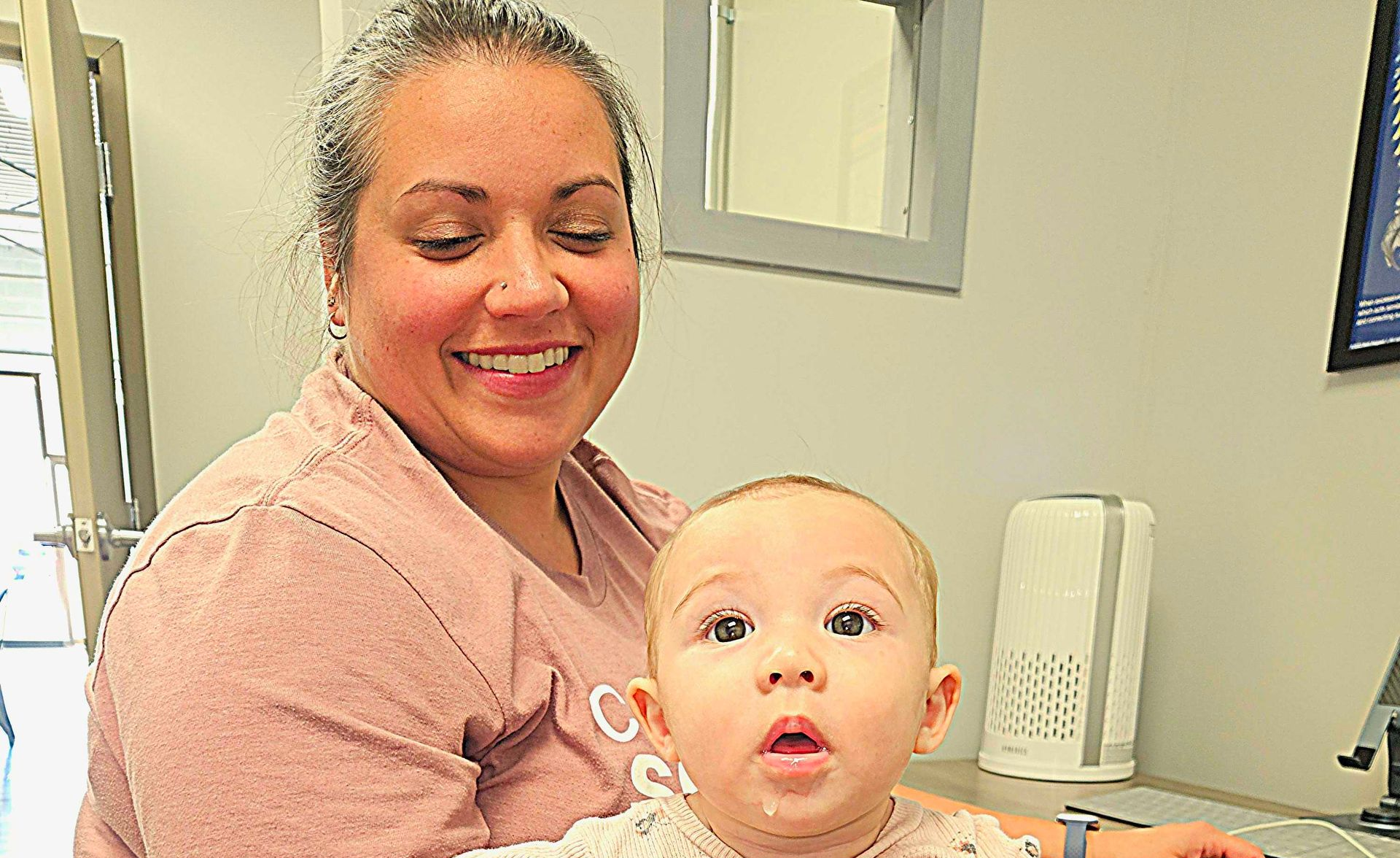 Woman holding a baby in a bright room, both looking at the camera and smiling.
