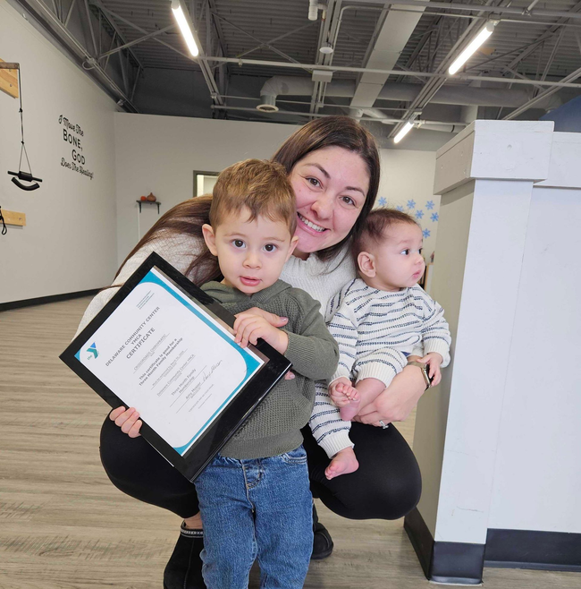 Woman crouching with two toddlers, holding a tablet in an office hallway.