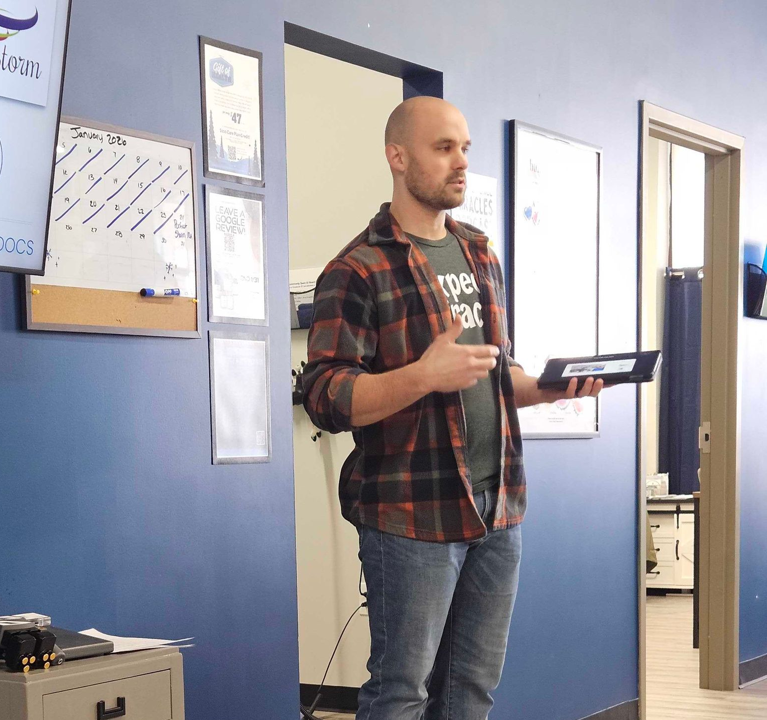 Person speaking in a classroom, holding a tablet and remote beside a blue wall with posters.