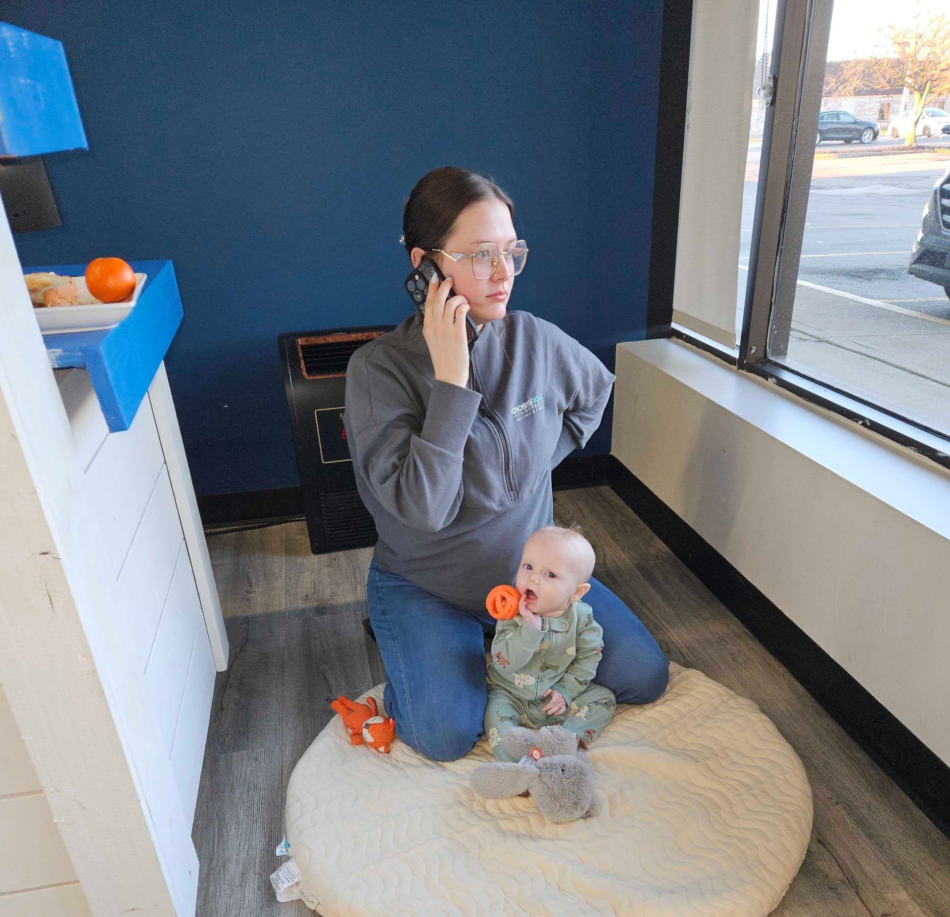 Woman kneels on a cushion, talking on a phone while holding a baby in a bright room by a window.