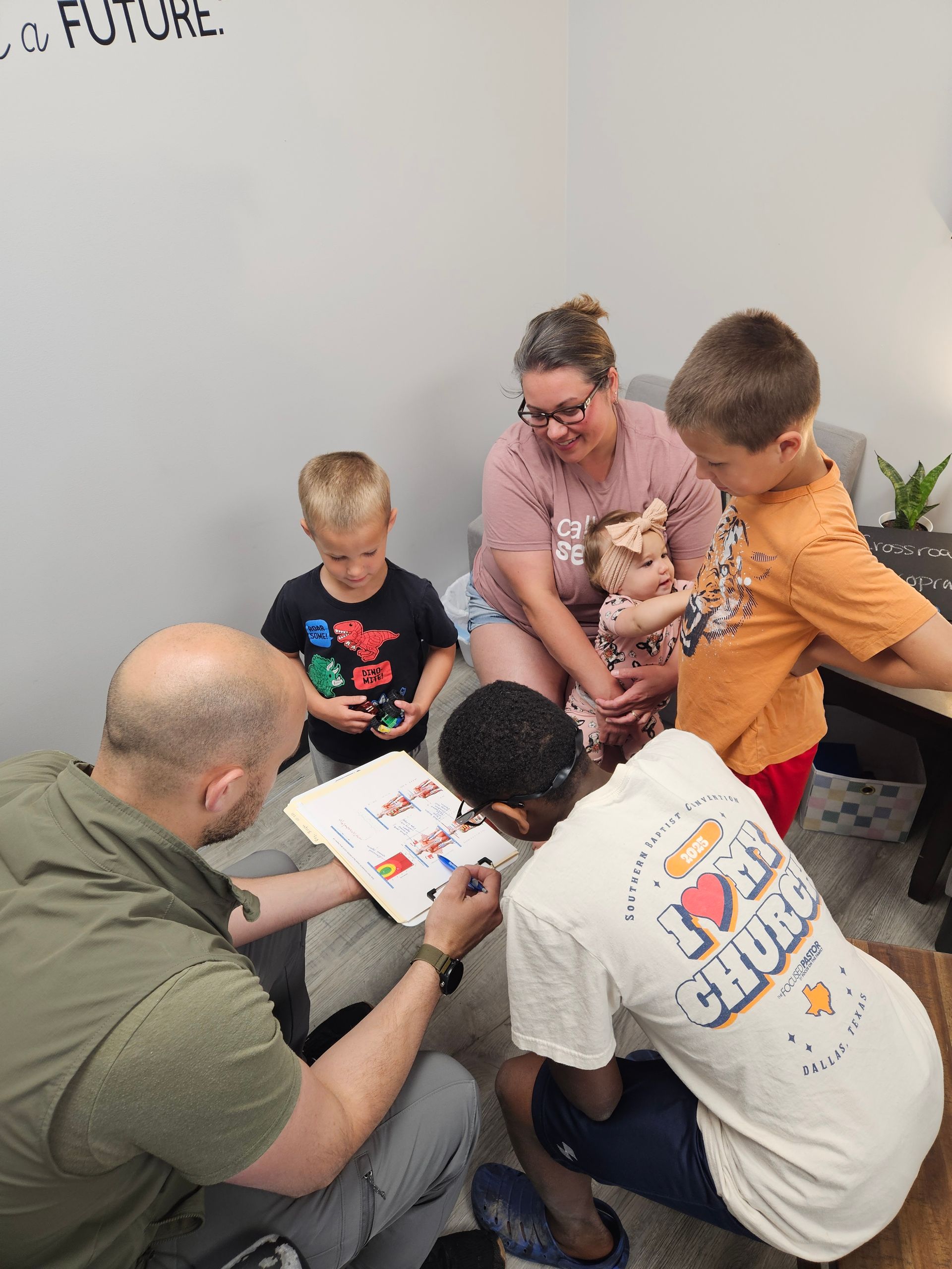 Group gathered around a book on the floor, with an adult guiding children in a bright room.