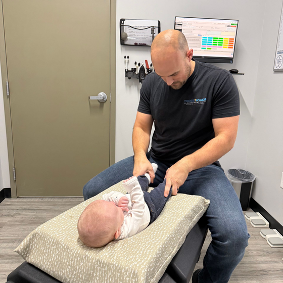 Man in a clinic gently stretches a baby's leg on a padded exam table.