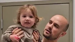 Man holding a smiling toddler in a beige room, both facing the camera.