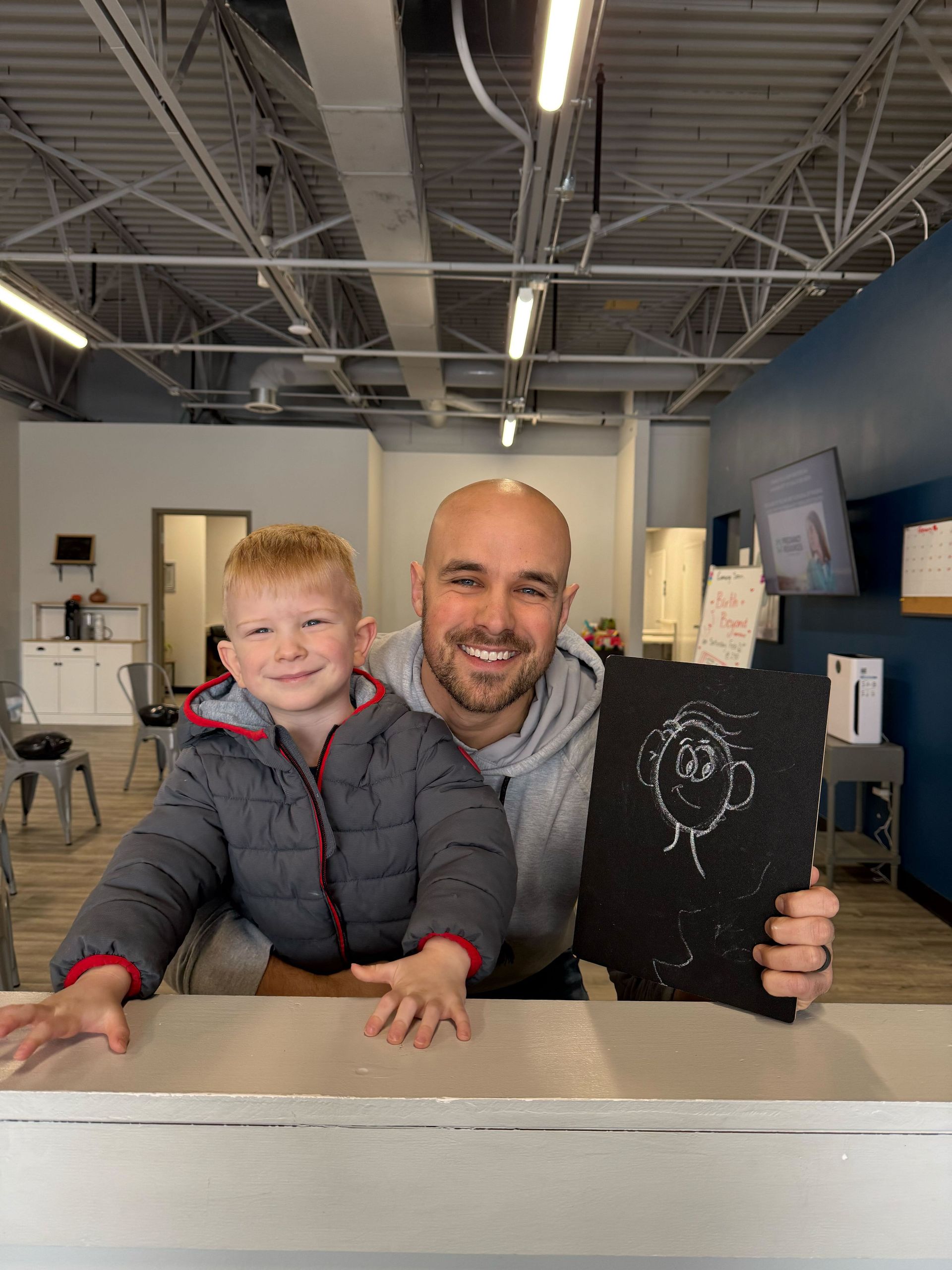 Two people smiling at a table in a bright office, holding a chalkboard with a lightbulb drawing.