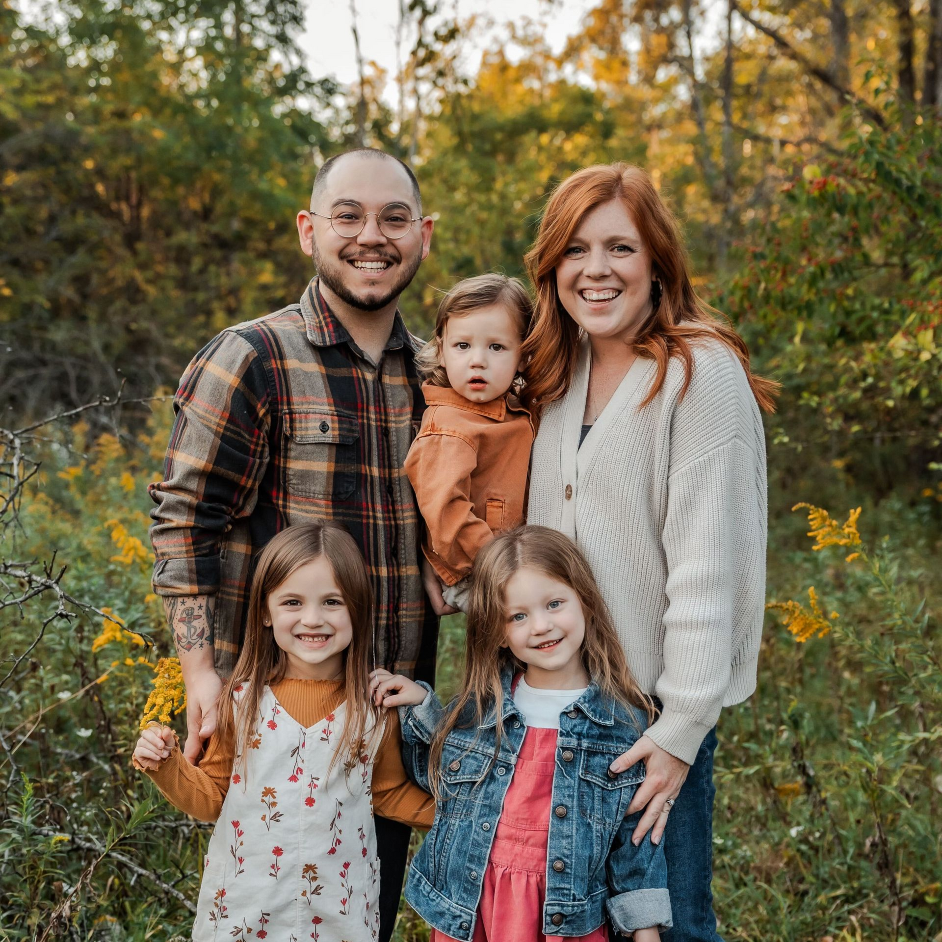 Family of five posing outdoors in a field, with autumn trees in the background.