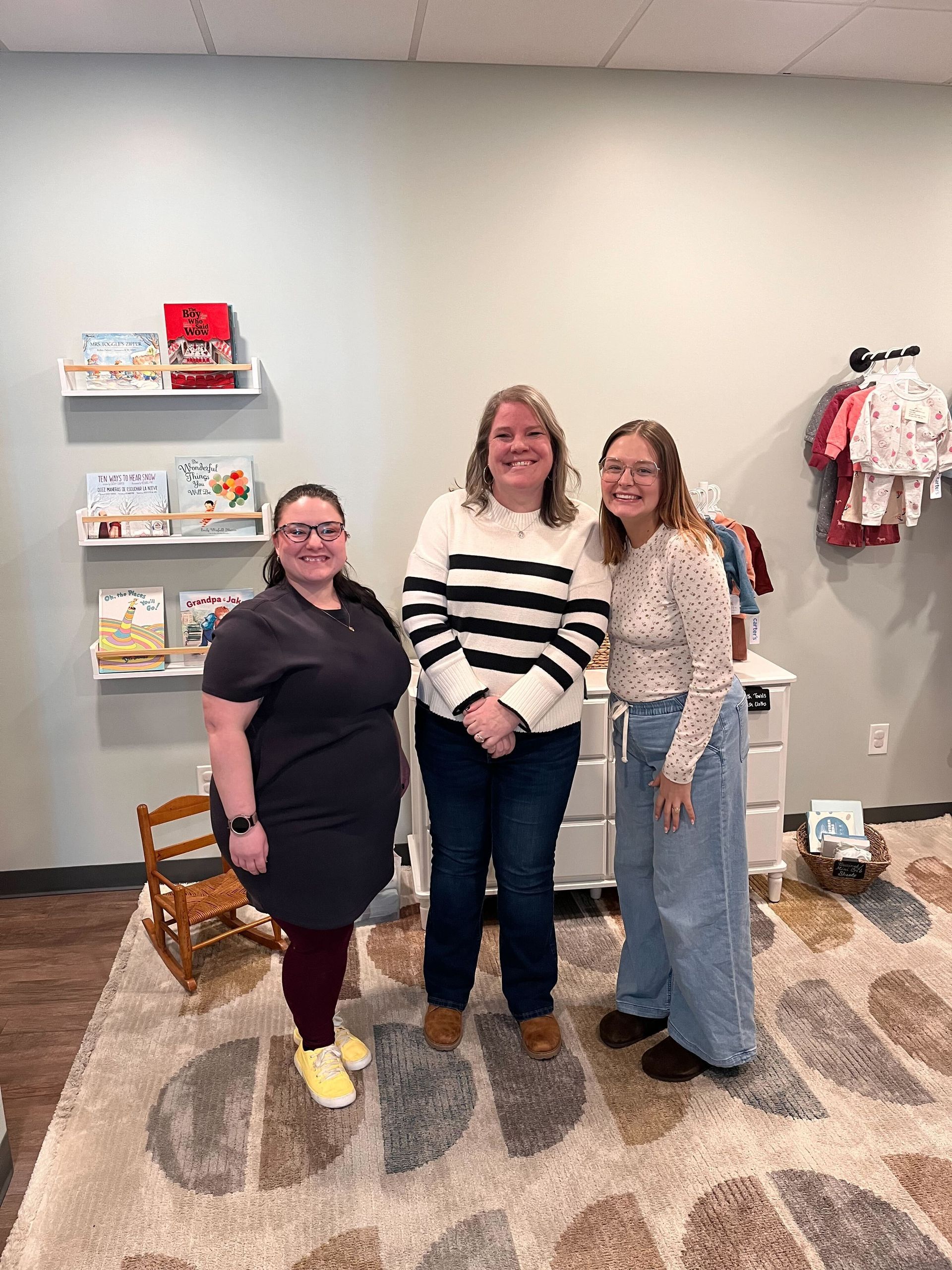 Three women standing together in a bright gallery, smiling near wall shelves and framed artwork.