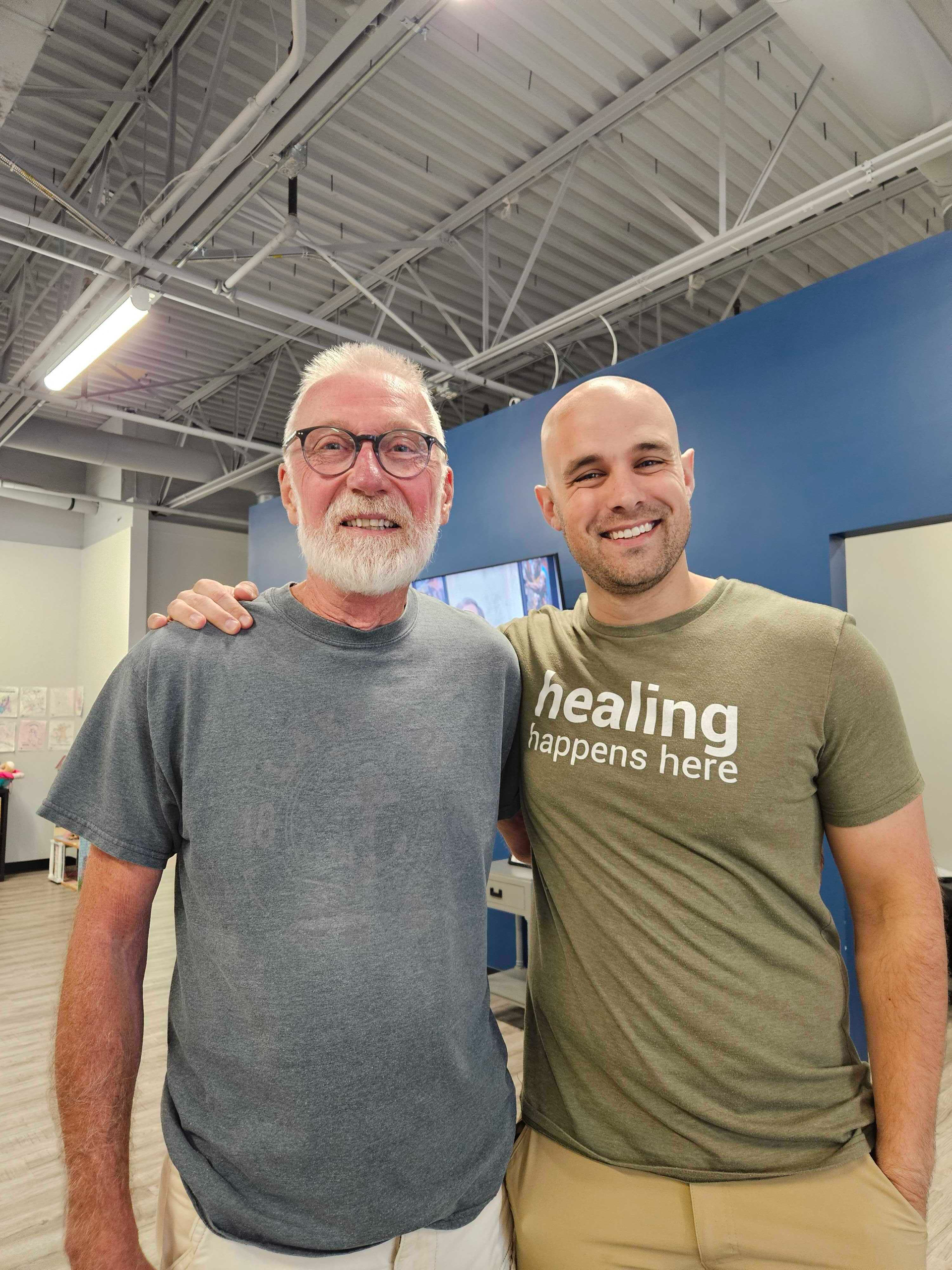Two smiling men standing together in a gym, one in a gray shirt and one in an olive “healing happens here” shirt.