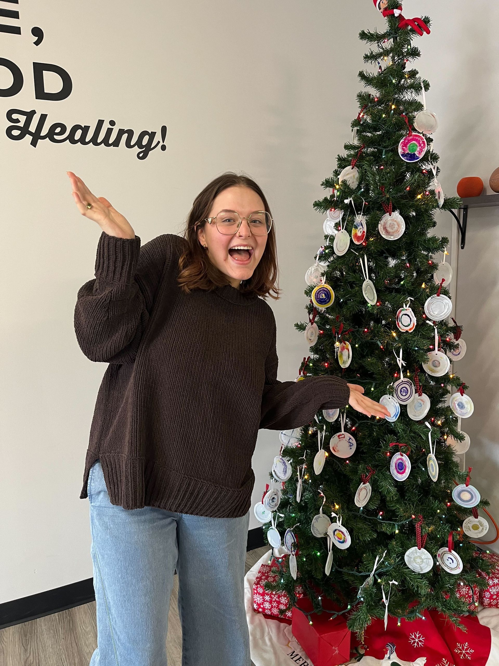 Woman posing beside a decorated Christmas tree indoors, smiling and gesturing toward it