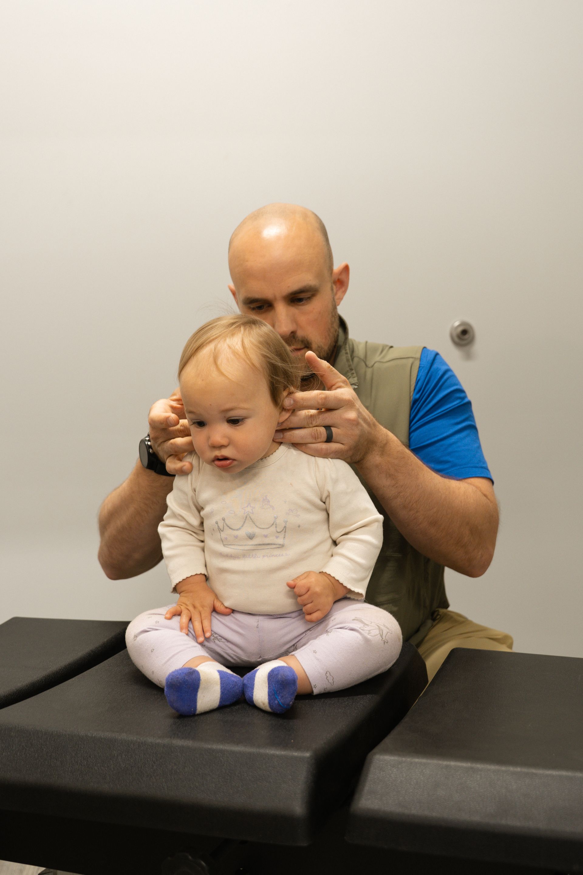 Adult examining a toddler’s ears in a clinic, with the child sitting on an exam table.