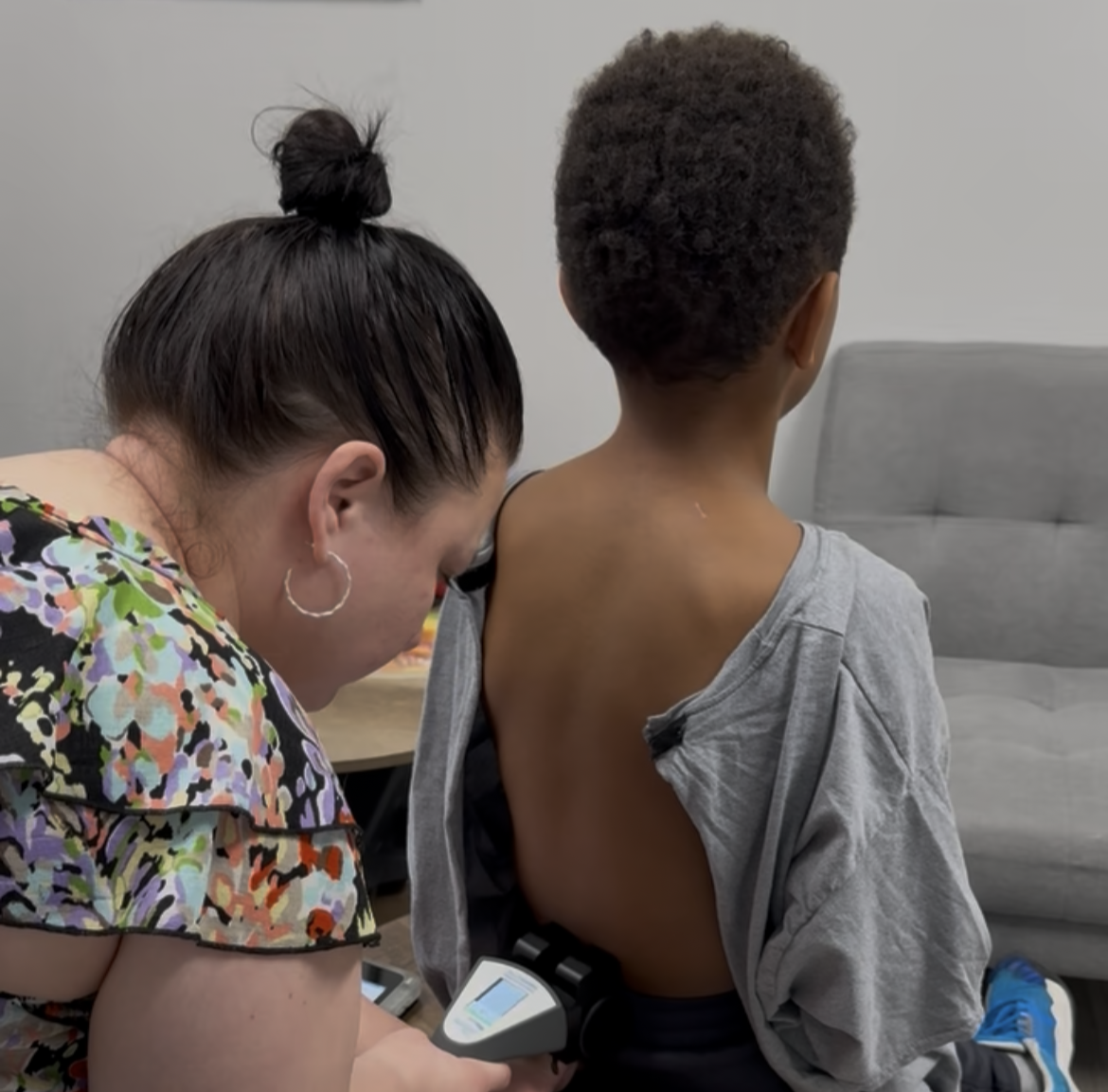 Woman using a handheld device on a child’s back in a living room, child seated facing away