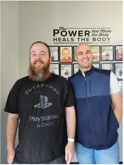 Two men standing indoors in front of a wall with the words “Power Heals the Body” and framed photos.