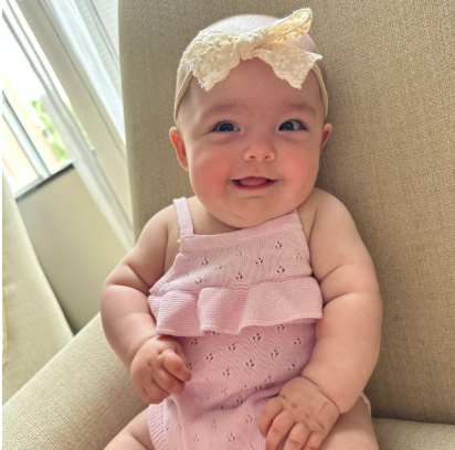 Smiling baby in a pink outfit with a cream bow, sitting on a beige chair by a sunlit window