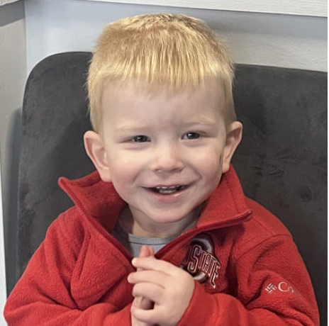 Smiling child in a red jacket sitting on a dark sofa.