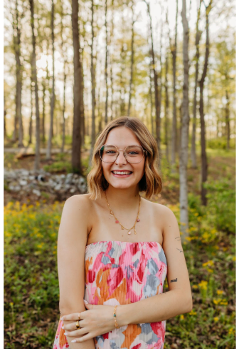 Smiling woman in a pink floral dress standing outdoors among trees in a wooded area.