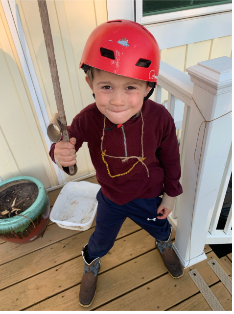 Child in a red helmet holding a tool, standing on a wooden porch beside a planter and white railing.