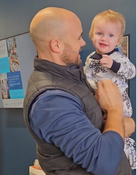 Man holding a smiling child indoors, both facing each other.