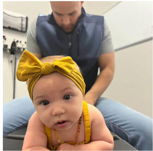 Baby on a mat wearing a yellow bow headband, with an adult behind during tummy time.