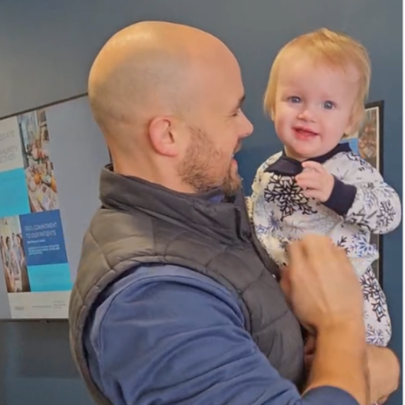 Man holding a smiling baby indoors, with office cubicles in the background.