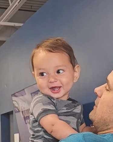 A smiling toddler in a gray shirt being held by an adult indoors against a blue wall.