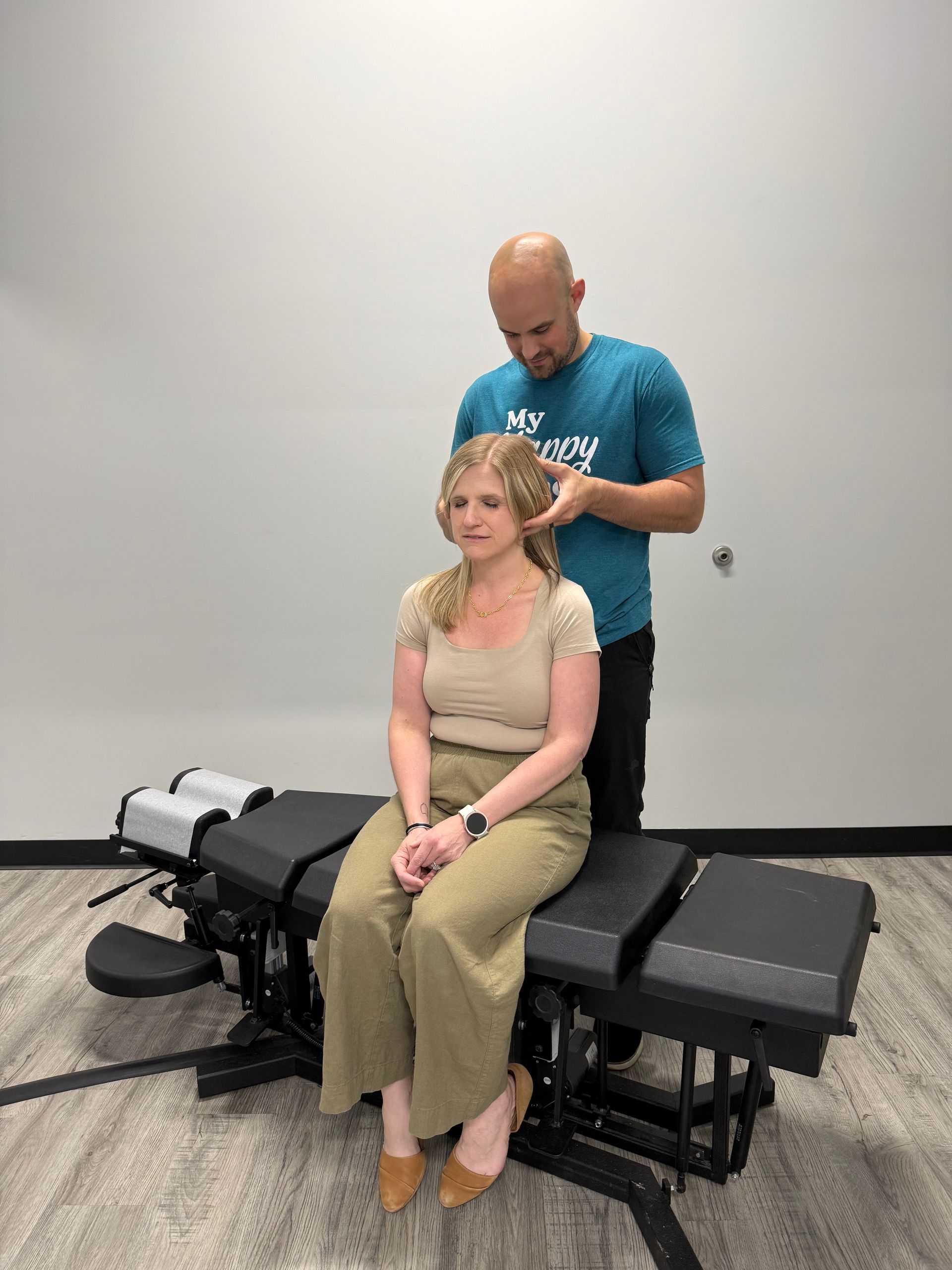 Person receiving a chiropractic adjustment while seated on an adjustment table in a clinic room.