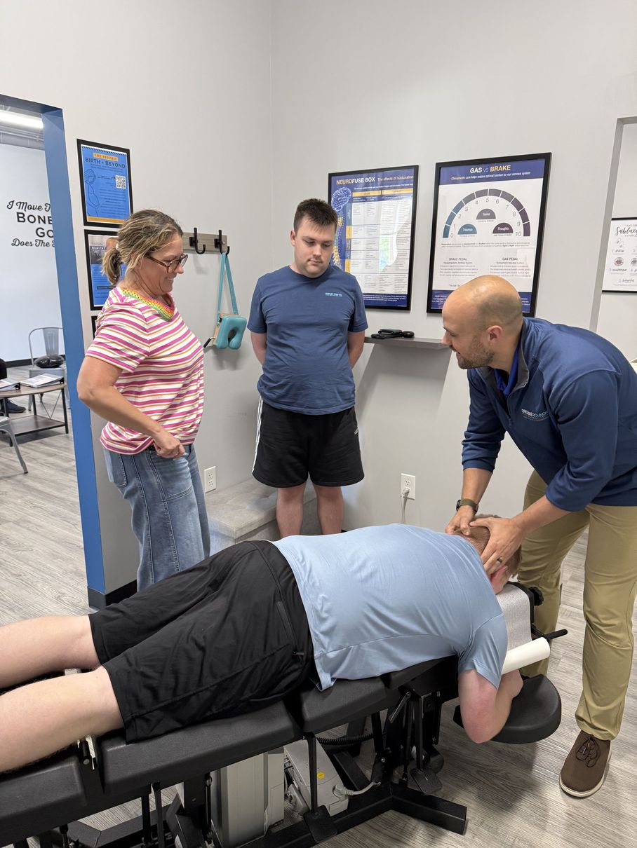 Trainer adjusts person on exam table while two others watch in a clinic room.