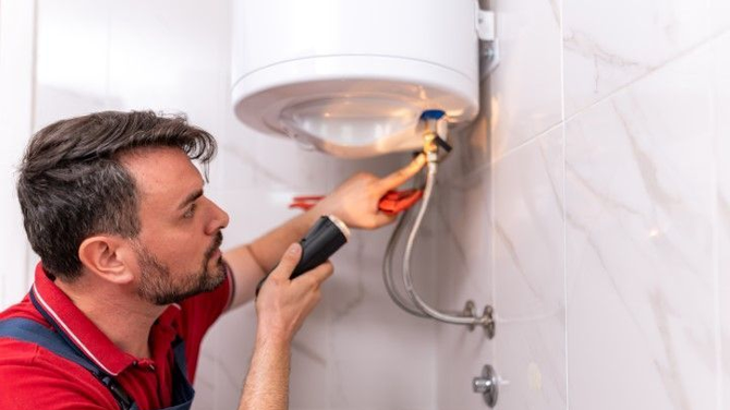Plumber repairing a wall-mounted water heater with a flashlight in a tiled bathroom