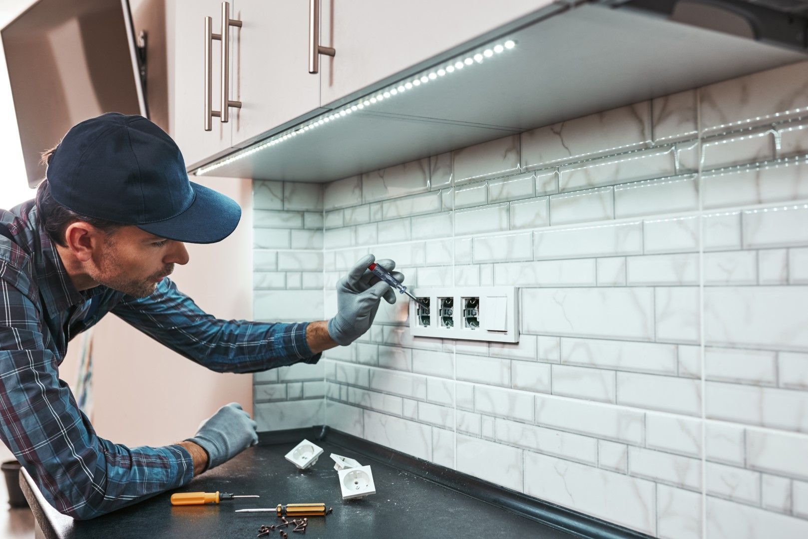 Electrician installing an outlet on a white tiled kitchen backsplash under a cabinet