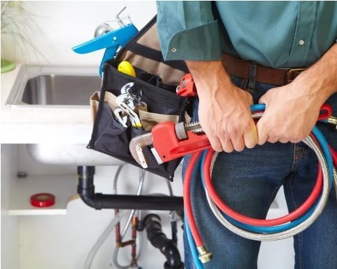 Plumber holding coiled red and blue hoses beside a sink and tool bag under a countertop