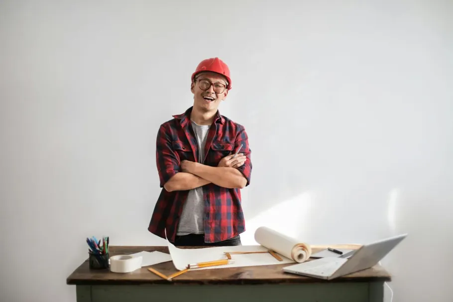 Person in a red hard hat standing behind a desk with blueprints and a laptop, smiling with arms crossed.