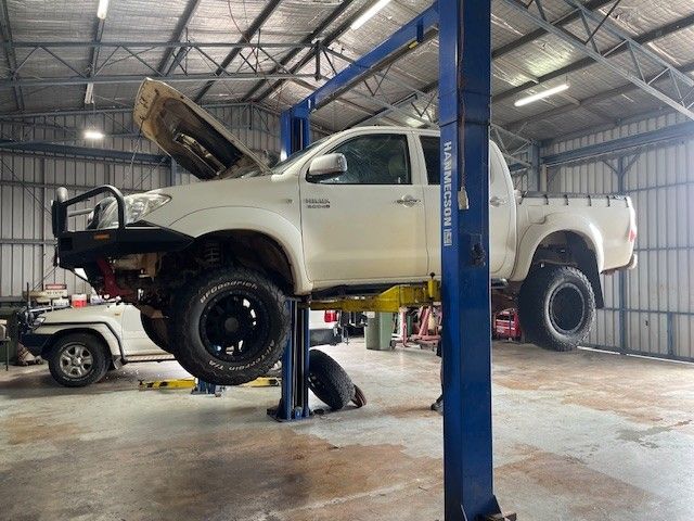 A white pickup truck on a car lift in a garage with a mechanic working underneath — JJE Automotive In Yarrawonga, NT