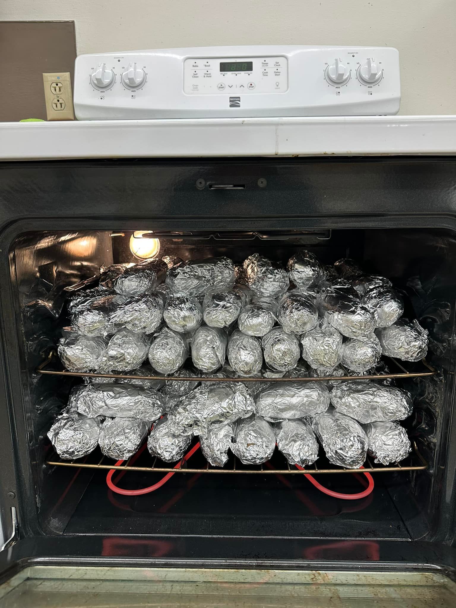 Oven with two racks, filled with foil-wrapped potatoes baking inside