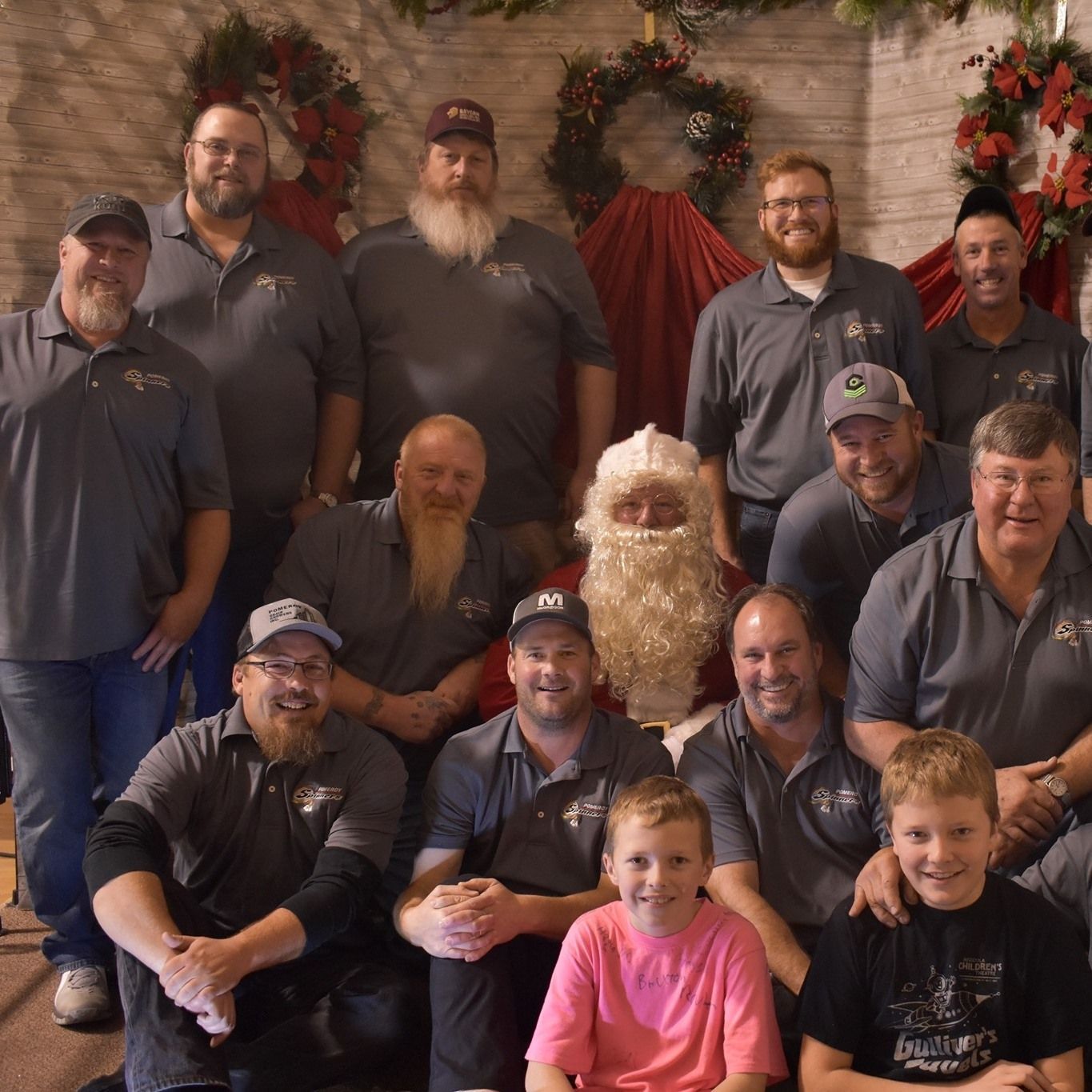 Group photo with Santa Claus. Men and two boys wear gray shirts. Christmas decorations in the background.