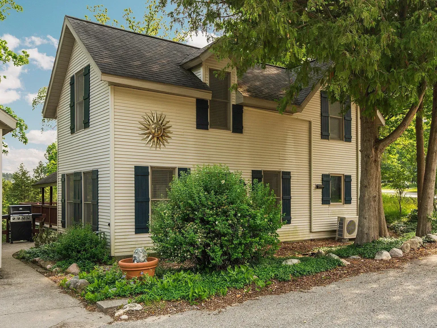 Two-story cream house with dark shutters, a sunburst wall ornament, and a tree-lined yard