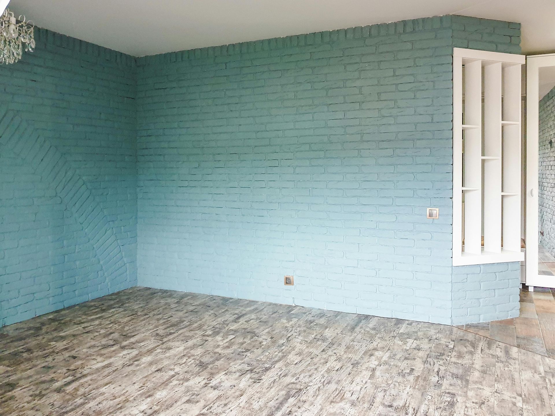 An empty room with light blue painted brick walls and gray patterned wood-look flooring, featuring a white window frame.