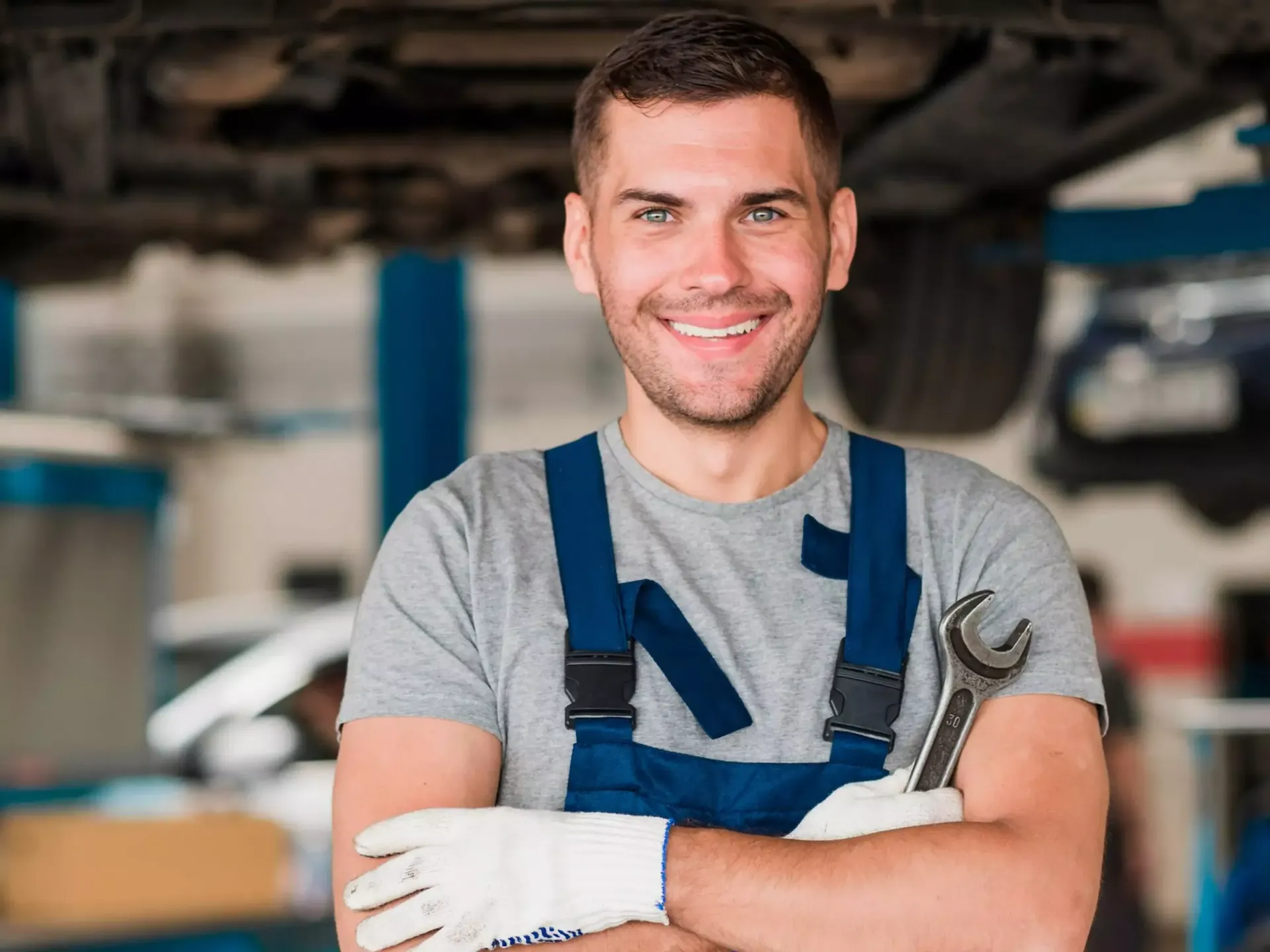 Um homem de macacão está segurando uma chave inglesa e sorrindo em uma garagem.