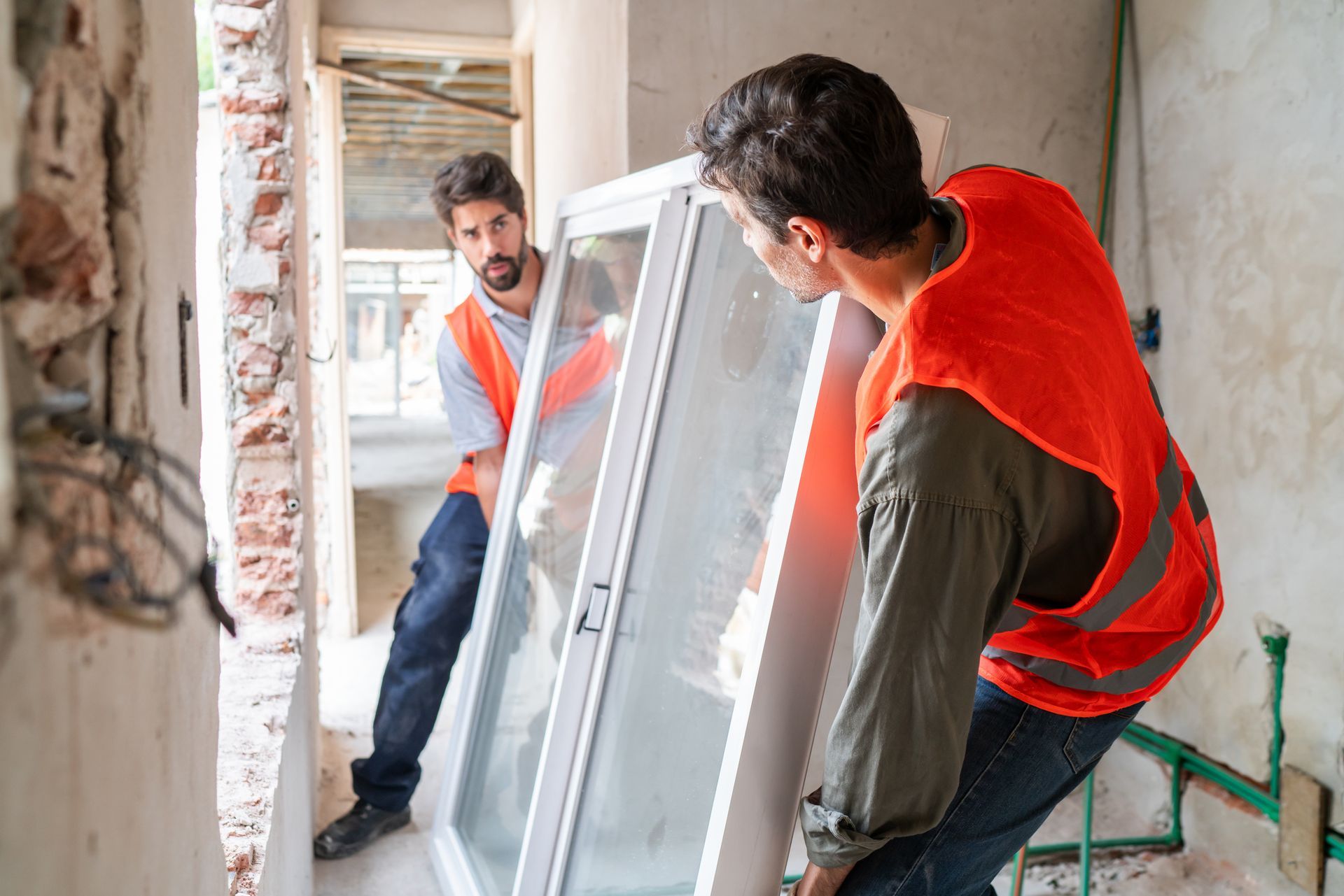 Two construction workers in orange vests are working on a residential window replacement at a house.