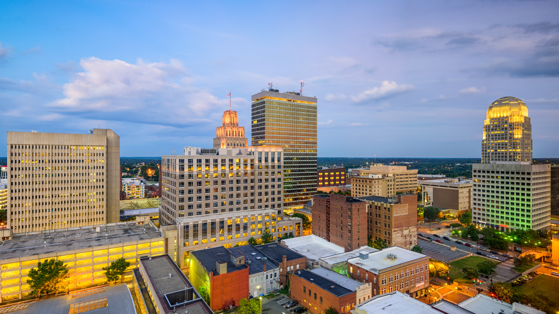 Dusk city skyline with lit office towers and buildings under a blue sky.