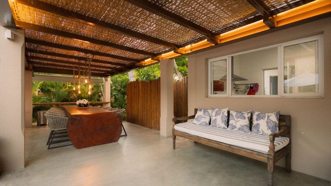 Covered patio with woven roof, wooden bench, and dining area beside a house window