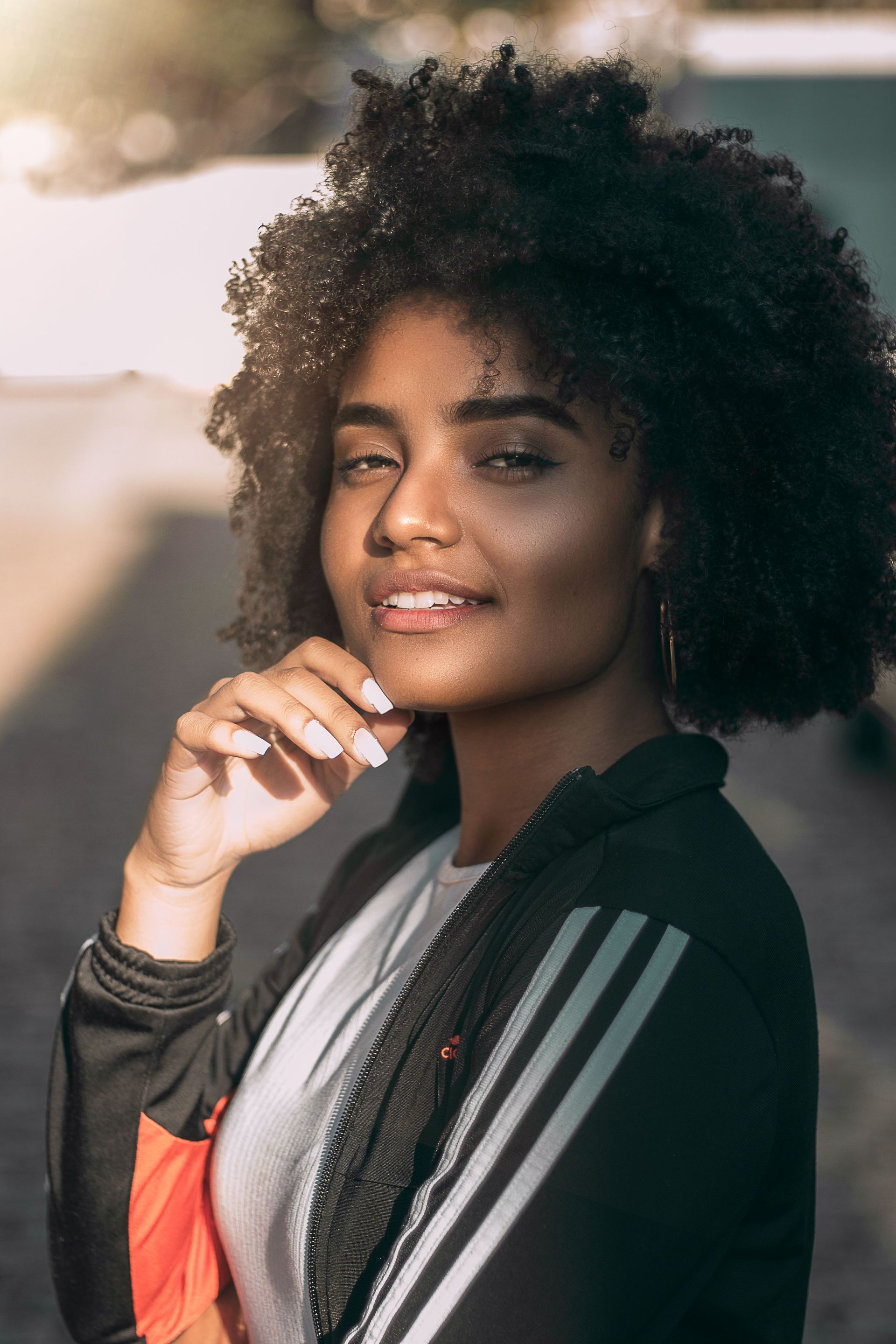 Portrait of a person with curly hair smiling, resting their hand on their chin, wearing a black jacket with white stripes.