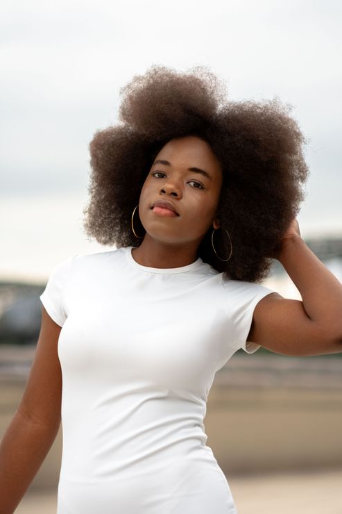Woman in a white shirt posing outdoors, hand in her hair, with a neutral expression.