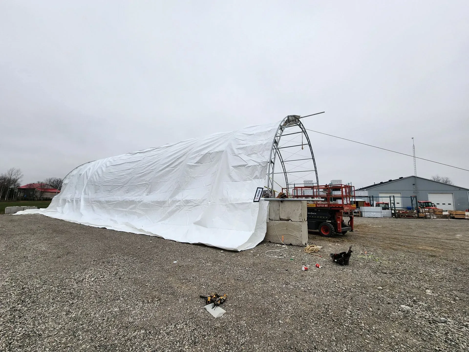 A white plastic-covered greenhouse frame stands on gravel beside a small tractor at a rural worksite.
