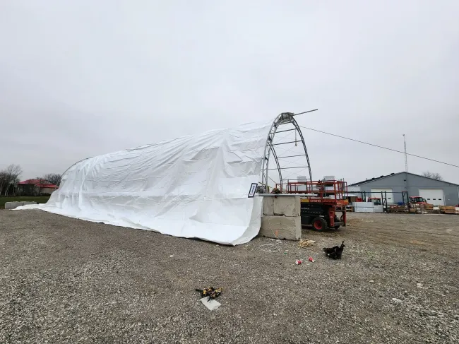 White tent-like tarp stretched over a framework on a gravel lot beside a utility vehicle and buildings