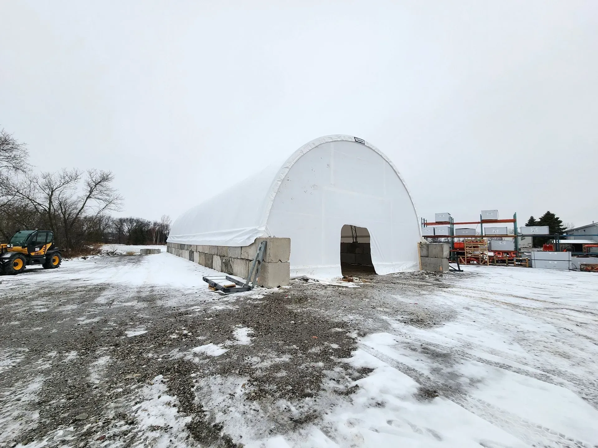 Snow-covered construction site with a white arched temporary building and muddy ground under an overcast sky