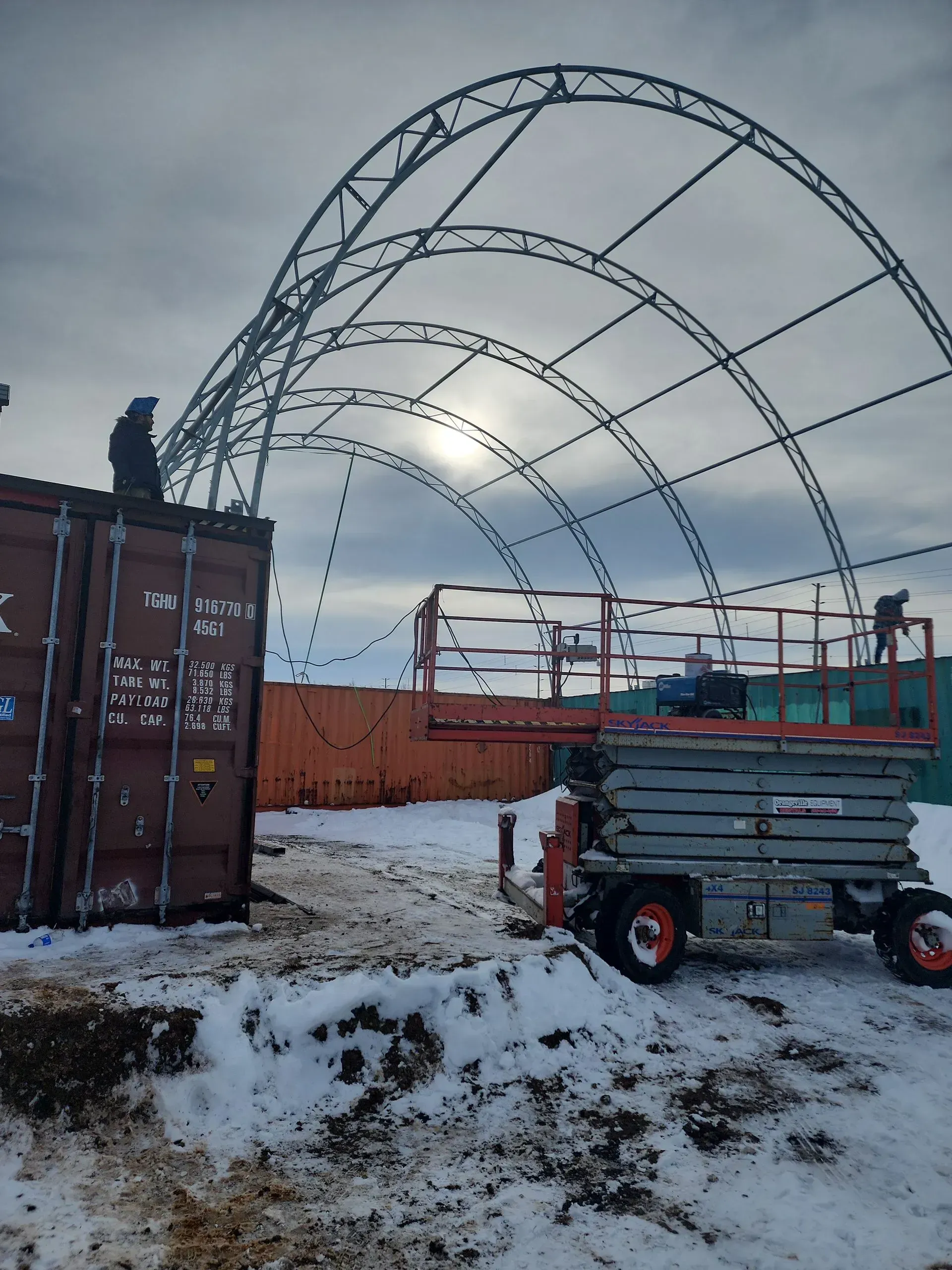 Snowy construction site with arched steel frame, shipping container, and utility trailer under a cloudy sky
