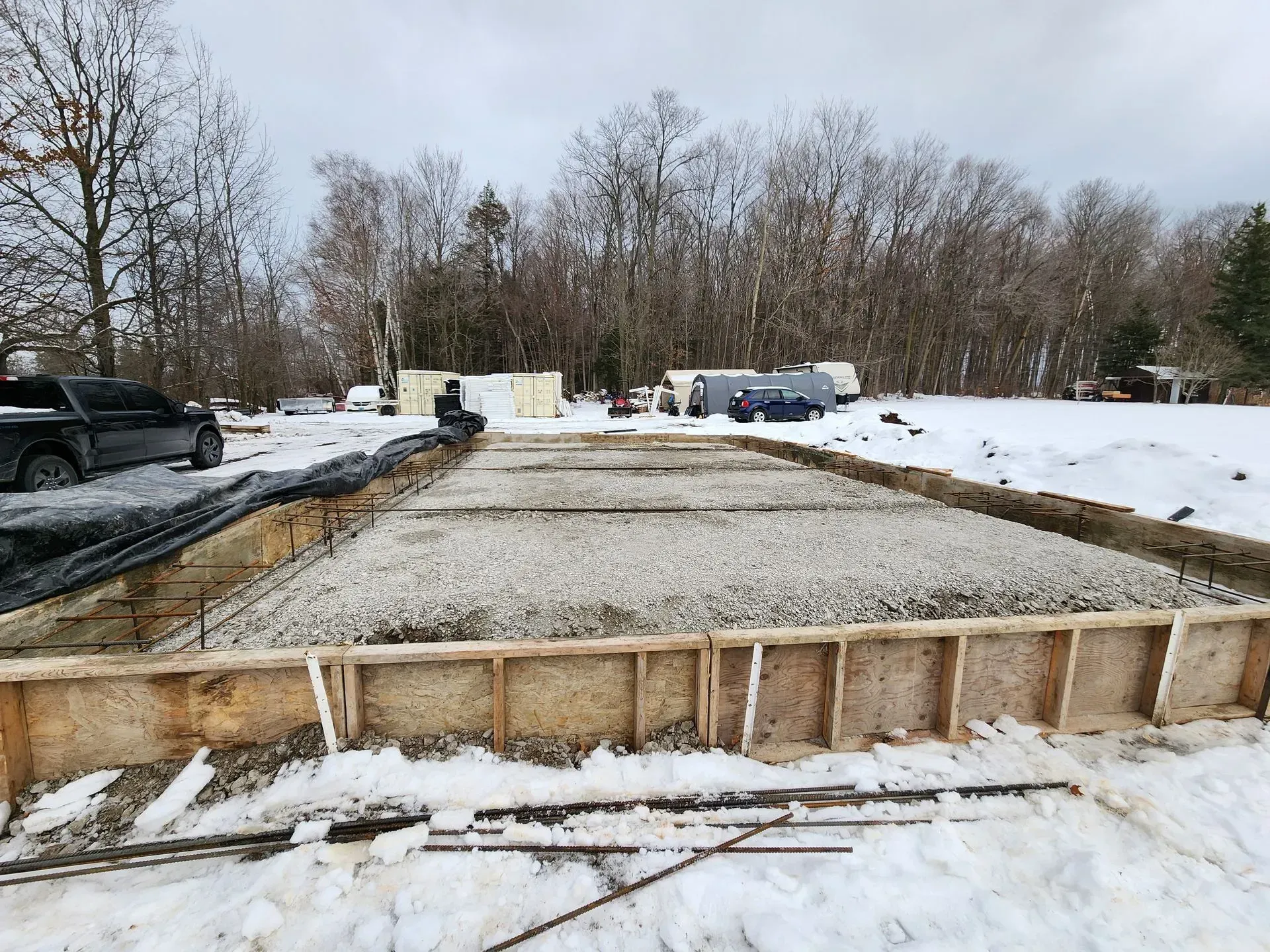 Snow-covered construction site with a gravel foundation and parked vehicles under overcast skies